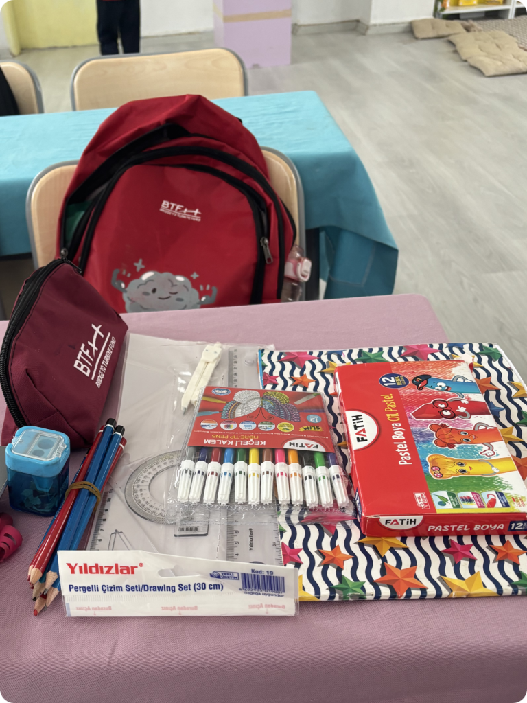 A red BTF backpack and matching pencil case displayed alongside notebooks, colored pencils, markers, pastels, and other essential school supplies provided through the 2026 Back to School Campaign.