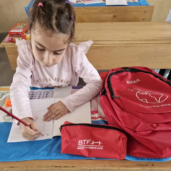 A student at Güzelyurt Elementary School in Adıyaman, Türkiye, practices writing beside her new red BTF backpack and matching pencil case.