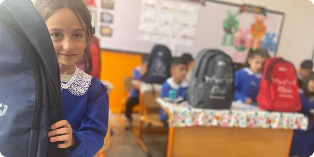 A young student in a blue school uniform holds a new backpack in a classroom, with other students and backpacks visible in the background.