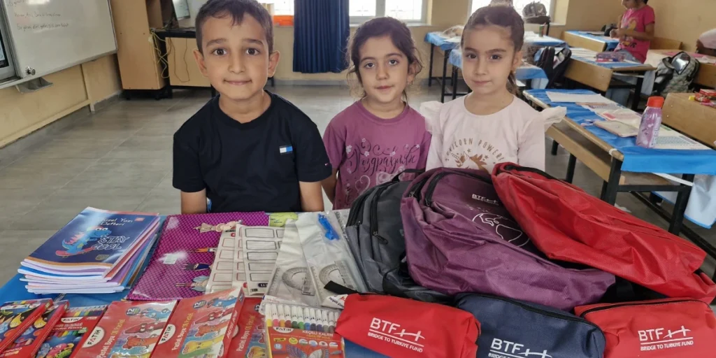 Three students at Güzelyurt Elementary School in Adıyaman, Türkiye, stand behind a table displaying BTF backpacks, pencil cases, notebooks, colored pencils, and other school supplies.
