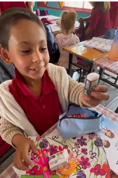 A young student at Haydarlar Elementary School in Hatay, Türkiye, smiles while examining new school supplies at her desk, including scissors, stationery, and a pencil case.