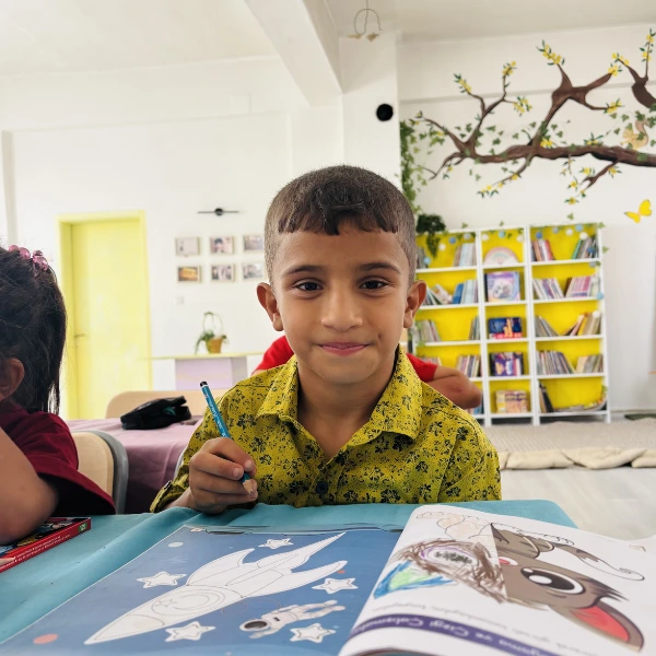 A student at Yastıca Elementary School in Kilis, Türkiye, smiles while holding a pencil over an open coloring book in his classroom.