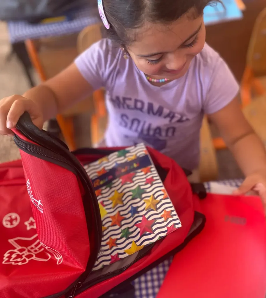 A young student in Ağrı, Türkiye, smiles while opening a red BTF backpack filled with notebooks and school supplies.