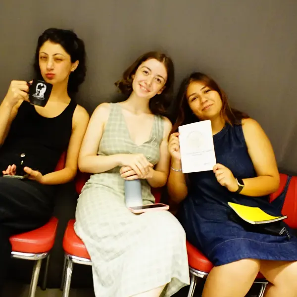 Three participants of the 2025 Nâzım Hikmet Summer Camp sit together indoors, with one holding a booklet and another holding a mug.