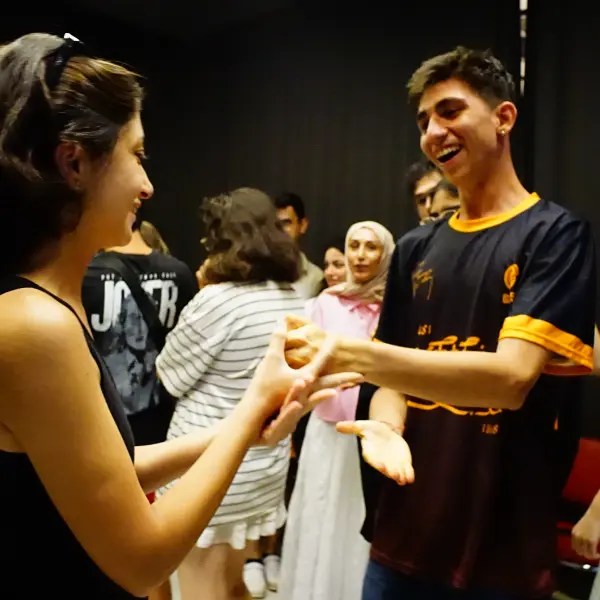Two participants smile and interact during a group activity at the 2025 Nâzım Hikmet Summer Camp, with other campers gathered in the background.